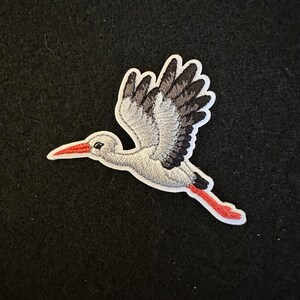 May include: A white and gray embroidered stork with black wings and a red beak and legs, flying in a black background.