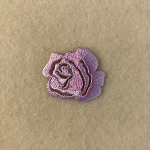 Puede incluir: Un pequeño parche bordado con forma de rosa de color morado claro. La rosa está delineada con un hilo morado más oscuro.