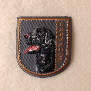 Puede incluir: Un parche bordado gris con un borde naranja. El parche presenta la cabeza de un Labrador Retriever negro con la lengua roja y la palabra "LABRADOR" escrita verticalmente en el lado derecho.