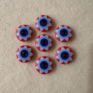 May include: Eight red and blue buttons with a flower design. The buttons have a white center with a blue circle and two black holes. The buttons are arranged in a circle.