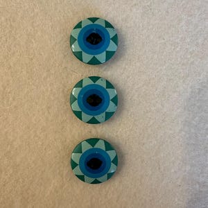 May include: Three round buttons with a blue and green eye design. The buttons have a white center and a black button in the center of the eye.