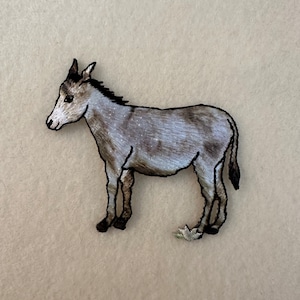 May include: A gray embroidered donkey patch with black outlines. The donkey is standing with its head turned to the left.