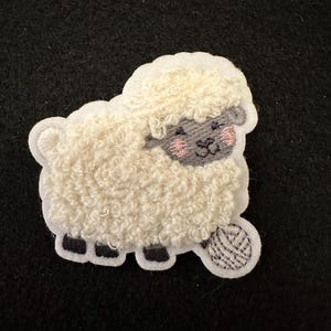May include: White fluffy sheep with a grey face and pink cheeks holding a grey yarn ball. The sheep is embroidered on a white background.