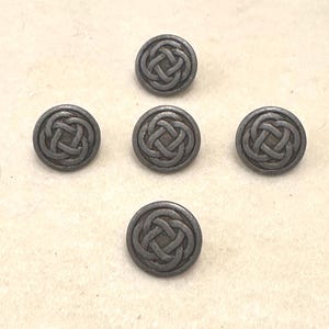 May include: Five antique silver-colored buttons with a Celtic knot design. The buttons are round with a raised, detailed knot pattern in the center. The buttons are arranged in a star-like pattern on a light beige surface.
