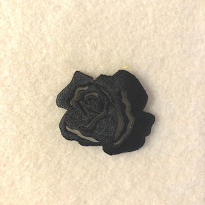 May include: A black embroidered rose patch featuring dense thread work that creates a textured floral design. The embroidery captures the layered petals of a blooming rose in a solid black colour, showcasing intricate stitching throughout the shape.