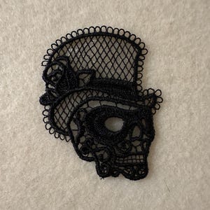 May include: Black lace skull applique with a top hat and rose detail. The skull has an open eye socket and detailed facial features. The applique is made of delicate, open-weave lace.