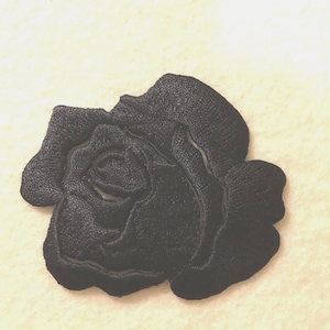 Puede incluir: Un parche bordado de una rosa negra con un trabajo de hilo denso que crea un diseño de pétalos texturizados y superpuestos. El bordado tiene forma de flor en floración con una espiral central y pétalos circundantes, con un acabado negro mate.