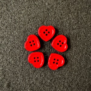 May include: Five red heart-shaped buttons with four black dots each. The buttons are arranged in a circle on a dark grey surface.