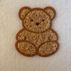 May include: A brown teddy bear patch with a textured, plush appearance. The bear has a dark brown outline and features, including small, round eyes and a nose. The patch is designed to be sewn or ironed onto fabric.