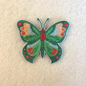 May include: Embroidered butterfly patch with green, red, orange, and blue details. The butterfly has a white border and is likely for decorative purposes, such as on clothing or accessories.