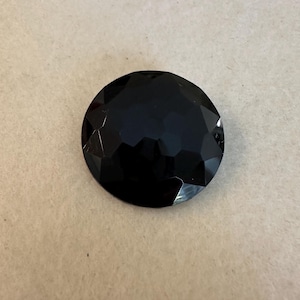 May include: A large, round, faceted black gemstone. The gem has a polished, reflective surface with multiple geometric facets, creating a sparkling effect. The stone is set against a neutral background.