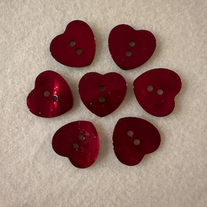 May include: Seven red heart-shaped buttons with two holes each. The buttons are shiny and have a metallic finish.