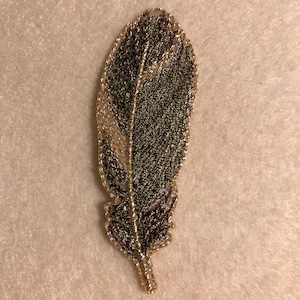 Puede incluir: Una pluma bordada en verde y dorado con un diseño detallado. La pluma es un adorno decorativo para manualidades y proyectos de costura.