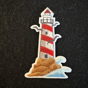 May include: Embroidered patch featuring a lighthouse with red and white stripes, a red roof, and a brown base. The lighthouse is set against a blue and white wave design, with a small bird in flight.