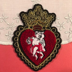 Puede incluir: Un parche en forma de corazón rojo con un borde dorado y un querubín en su interior. El corazón está coronado con una corona dorada con cuentas negras.