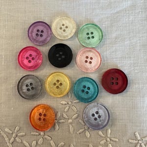 May include: A collection of eleven round, four-hole buttons in various translucent colors including purple, cream, green, pink, black, yellow, teal, red, orange, gray, and light purple, arranged in a scattered pattern on a textured fabric surface.