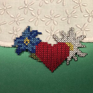 Könnte beinhalten: Kreuzstich-Blumen- und herzförmige Aufnäher in Blau, Rot, Weiß und Gelb. Die Aufnäher befinden sich auf einer grünen Oberfläche, mit einem weißen Stoffhintergrund mit gestickten Blumen.