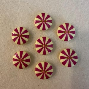 May include: Seven round buttons with a cream-colored base and a vibrant pink striped design radiating from the center. Each button has two small holes for sewing. The buttons are arranged on a light-colored surface.