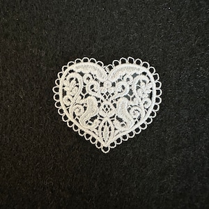 Puede incluir: Aplique de encaje blanco en forma de corazón con intrincados diseños florales. El aplique está sobre un fondo negro.