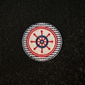 May include: A white, red, and blue embroidered patch with a nautical wheel design. The patch has a white background with a red circle around the wheel and a blue and white striped border.