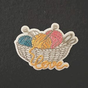 May include: Embroidered patch showing a basket of yarn balls in teal, yellow, and pink, with knitting needles. The word "love" is embroidered in yellow cursive below the basket. The patch has a white border.