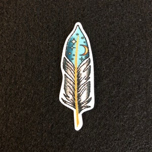 May include: A blue and gray embroidered feather with a crescent moon and stars in the center. The feather is outlined in gold.