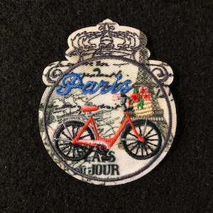 May include: A white and grey embroidered patch featuring a red bicycle, a basket of flowers, and the word "Paris" in blue lettering. The patch has a crown at the top and the words "Bon Jour" at the bottom.