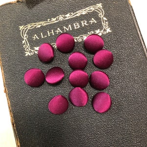 May include: A collection of 10 burgundy satin buttons, arranged on a dark brown book with the word "ALHAMBRA" embossed on the cover.