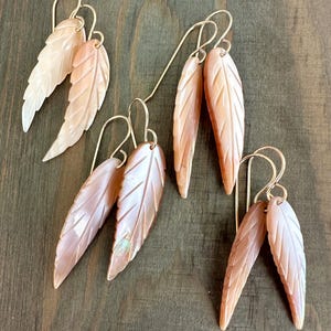 May include: Six pairs of leaf-shaped earrings with gold-colored hooks. The earrings are made of a light peach-colored material with carved details, resembling feathers. The earrings are arranged on a dark wood surface.