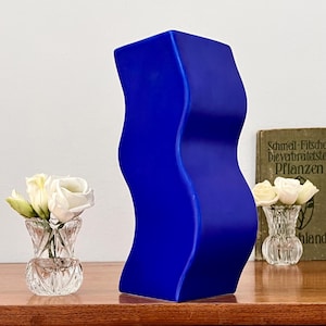 Op de afbeelding: Een levendige blauwe, golvende vaas. Twee kleine glazen vazen ​​bevatten witte rozen, geplaatst op een houten oppervlak. Een vintage boek is zichtbaar op de achtergrond. De vaas heeft een uniek, modern ontwerp.