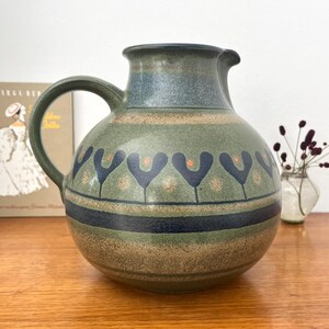 Könnte beinhalten: Ein runder Keramikkrug mit einer gedeckten grünen Basis und einem horizontalen Band aus dunkelblauen, stilisierten Blumenmotiven. Das Design umfasst gesprenkelte Texturen, einen breiten Henkel und einen ausgestellten Rand mit einem dunkelblauen Streifen.