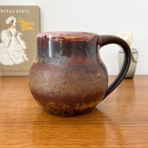 Könnte beinhalten: Eine handgefertigte Keramiktasse mit einem dunkelbraunen Henkel und einem abgerundeten Körper. Die Tasse hat ein strukturiertes, erdiges Design mit Brauntönen, Rot und Gelb. Die Tasse steht auf einer Holzoberfläche.