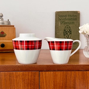 Può includere: Due contenitori in ceramica bianca con disegni a quadri rossi e neri. Il contenitore a sinistra è una zuccheriera e il contenitore a destra è un creamer. Entrambi i contenitori sono su una superficie di legno.