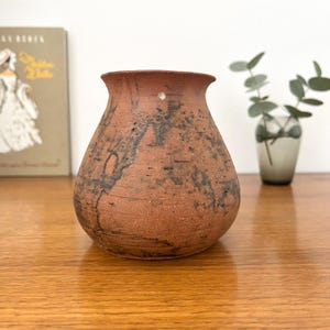 Könnte beinhalten: Eine terrakottafarbene Keramikvase mit einem breiten, bauchigen Körper und einem ausgestellten Rand. Die Oberfläche hat ein strukturiertes, mattes Finish mit dunklen, unregelmäßigen schwarzen Flecken und Streifen, die das Gefäß umziehen.