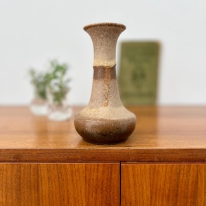 Può includere: Un vaso in ceramica marrone con superficie strutturata. Il vaso ha una base arrotondata, un collo sottile e un bordo svasato. Il colore passa da un marrone scuro alla base a un beige più chiaro verso l'alto. Il vaso è esposto su una superficie di legno.