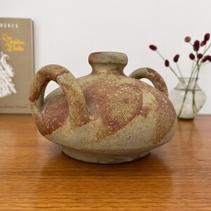 Könnte beinhalten: Ein rundes Keramikgefäß mit zwei bogenförmigen Henkeln und einem kurzen, schmalen Hals. Die Oberfläche weist ein gesprenkeltes, strukturiertes Finish in erdigen Tönen von Beige, Hellbraun und Rotbraun auf dem gesamten runden Körper auf.