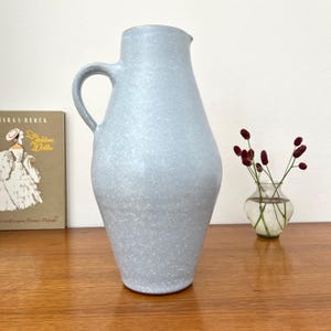 Puede incluir: Una jarra de cerámica alta de color azul claro con un acabado blanco moteado, que presenta un asa curva y un cuerpo ancho y cónico. La jarra tiene una textura suave y mate, con un diseño funcional que incluye un pico vertedor en el borde superior.