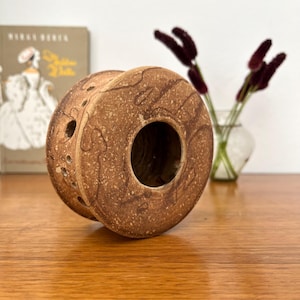 May include: A circular, speckled brown ceramic object with a hollow centre and a series of small, evenly spaced circular perforations along its side. The surface features a textured, mottled finish with darker brown swirling patterns throughout.