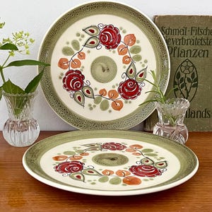 Könnte beinhalten: Zwei Vintage-Keramikplatten mit floralem Design. Die Teller haben einen cremefarbenen Hintergrund, einen grünen Rand und ein zentrales Blumenmuster mit roten Rosen und grünen Blättern. Ein Buch mit deutschem Text ist sichtbar.