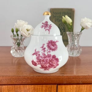 Może przedstawiać: Biała cukiernica z porcelany z pokrywką, ozdobiona kwiatowym wzorem w odcieniach różu i złota. Cukiernica ma okrągły kształt i delikatny wzór kwiatowy.