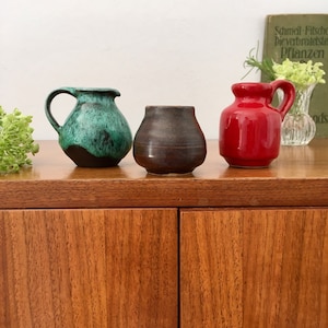 Peut inclure: Trois petits vases en céramique, un vert, un brun et un rouge, sont disposés sur une surface en bois. Les vases sont de formes et de tailles différentes.