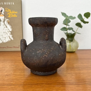 Könnte beinhalten: Kleine, dunkelbraune Keramikvase mit zwei Henkeln und einer gesprenkelten Textur. Die Vase hat einen runden Körper und einen ausgestellten Rand. Sie steht auf einer Holzoberfläche.