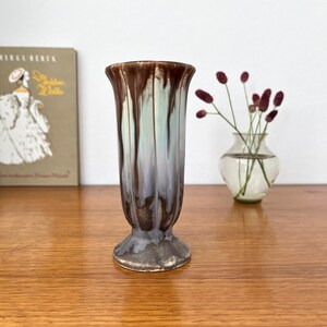Könnte beinhalten: Eine Keramikvase mit geriffeltem Sockeldesign und einer vertikalen Tropfglasur in den Farben Dunkelbraun, Hellblau und Weiß. Die Vase hat einen ausgestellten Rand und eine glatte, glänzende Oberfläche und steht aufrecht als einzelnes Objekt.
