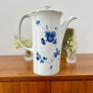Könnte beinhalten: Eine weiße Porzellan-Kaffeekanne mit blauem Blumendesign. Die Kanne hat einen gebogenen Griff, einen Ausguss und einen Deckel. Das florale Muster umfasst Blütengruppen und zarte Stiele. Die Kanne steht auf einer Holzoberfläche.