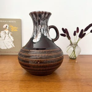 May include: A ceramic pitcher vase with a dark brown glazed body featuring a textured, repeating geometric pattern around the base. The neck has a white drip glaze effect over a dark brown background, and a single handle is attached to the side.
