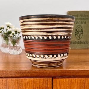 Puede incluir: Una maceta de cerámica con un diseño a rayas marrón, crema y rojo. La maceta presenta una banda de puntos blancos y está colocada sobre una superficie de madera. A la izquierda se ve un pequeño jarrón con flores blancas.