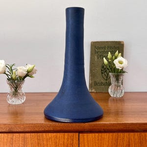 Könnte beinhalten: Eine hohe, dunkelblaue Keramikvase mit einem ausgestellten Fuß. Die Vase steht auf einer Holzoberfläche mit einem Buch und einer kleinen Vase mit weißen Blumen.