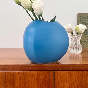 May include: A blue ceramic vase with a rounded, organic shape sits on a wooden surface. White flowers are arranged in the vase.