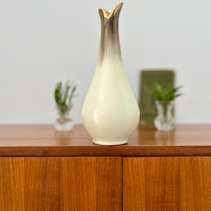 Könnte beinhalten: Eine hohe, cremefarbene Vase mit einem ausgestellten, goldfarbenen Oberteil und einem braunen Farbverlauf. Die Vase steht auf einem Holzschrank. Zwei kleine Glasvasen mit Grünzeug befinden sich im Hintergrund.