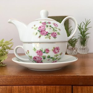 Könnte beinhalten: Weißes Teeset mit floralem Design mit rosa und weißen Rosen. Die Teekanne und die Tasse sind übereinander gestapelt und das Set steht auf einer Holzoberfläche.